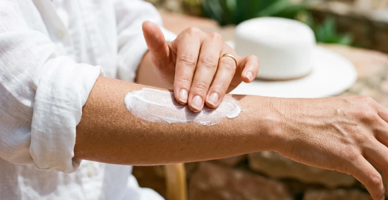 Mains féminines appliquant de la crème solaire blanche sur l'avant-bras dans un environnement lumineux.